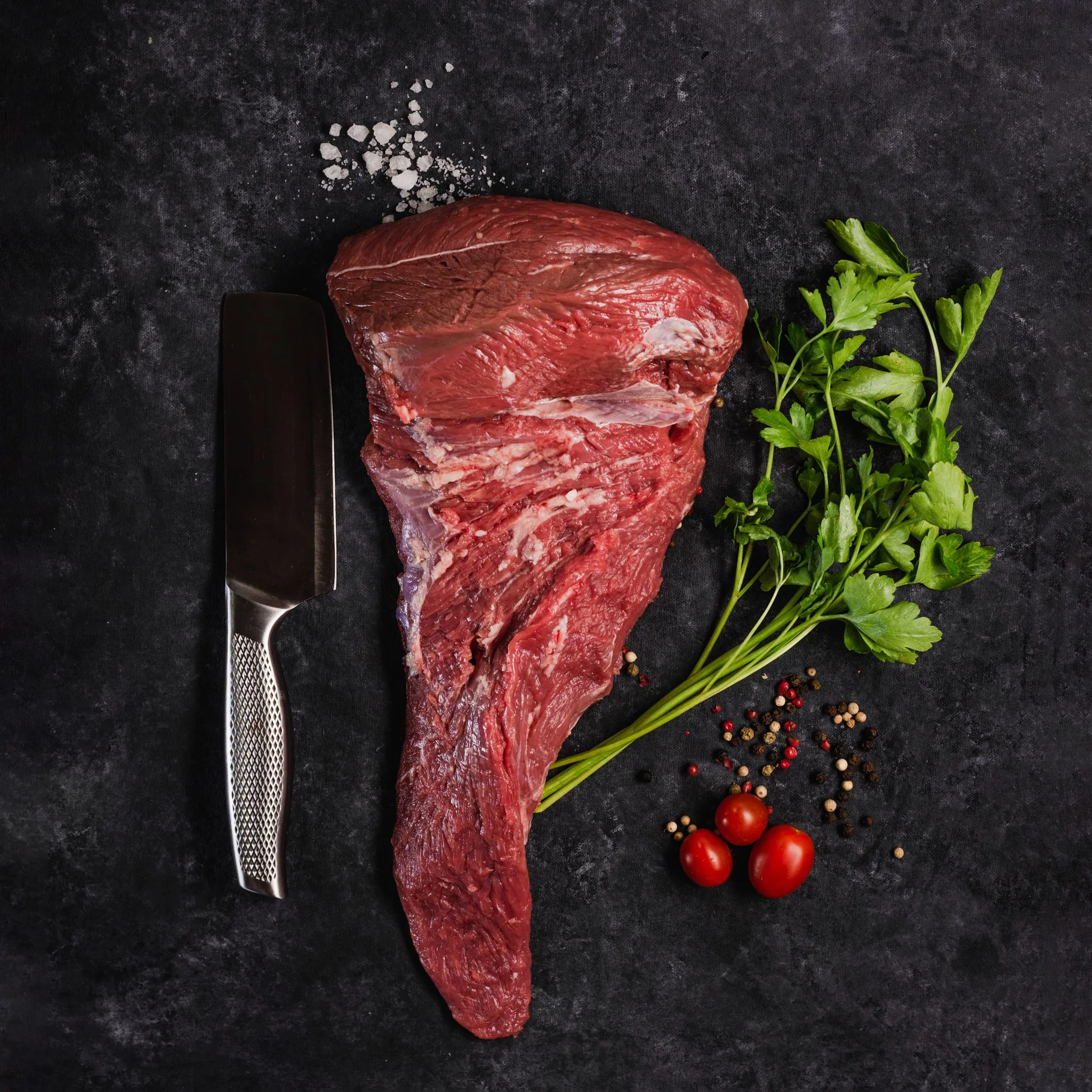 A raw Argentine Angus picanha with a cleaver, parsley and peppercorns on dark stone